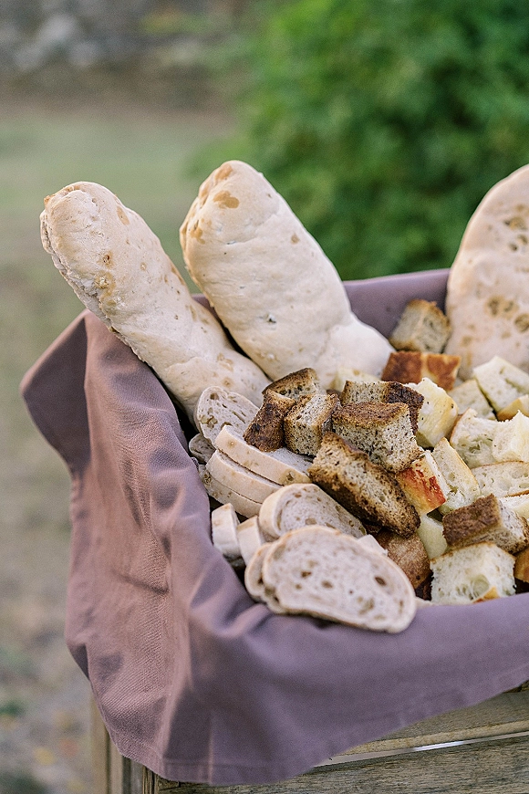 Wedding bread basket lined with a linen napkin holding baguettes, sliced bread, and bread cubes, set against blurred outdoor greenery
