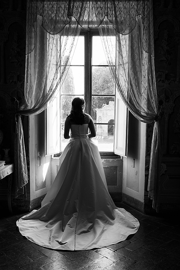 Bridal portrait of a bride at window, back view showing a strapless gown with long train and button details beside lace curtains