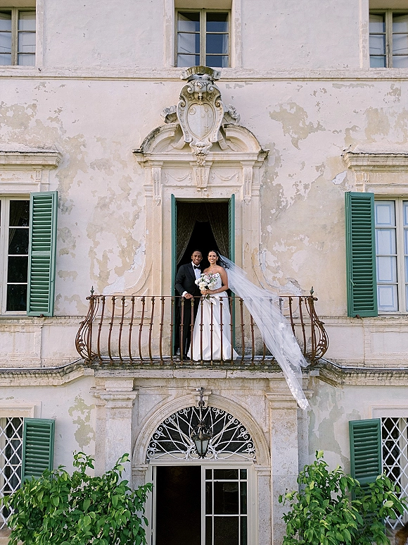 Couple portrait of bride and groom balcony pose, her long veil blowing as she holds a bouquet beside him in a black tuxedo by a historic stucco facade