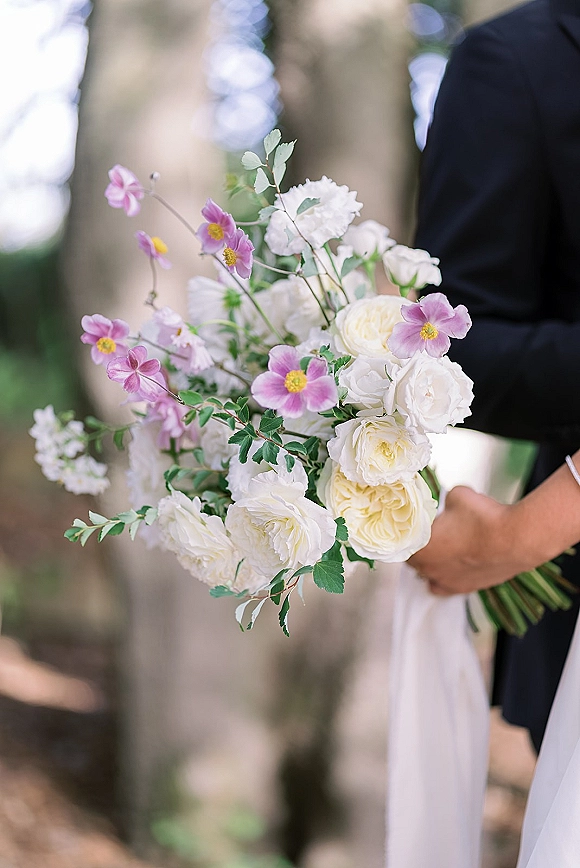 Bridal bouquet of white roses and pink anemones with greenery, held by the bride beside groom in an outdoor garden setting
