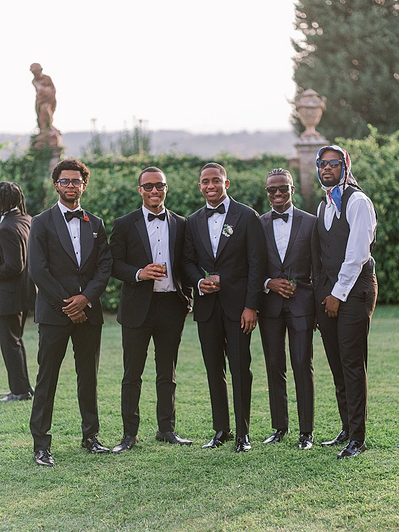 Groomsmen portrait of men in tuxedos wearing sunglasses and holding cocktail glasses, lined up on a lawn with hedges and statues behind