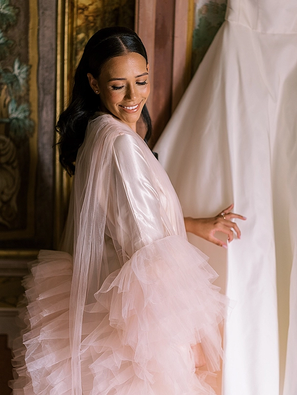 Bridal portrait of a bride getting ready in a tulle robe and veil, looking down at her ring beside a hanging gown and ornate wall art