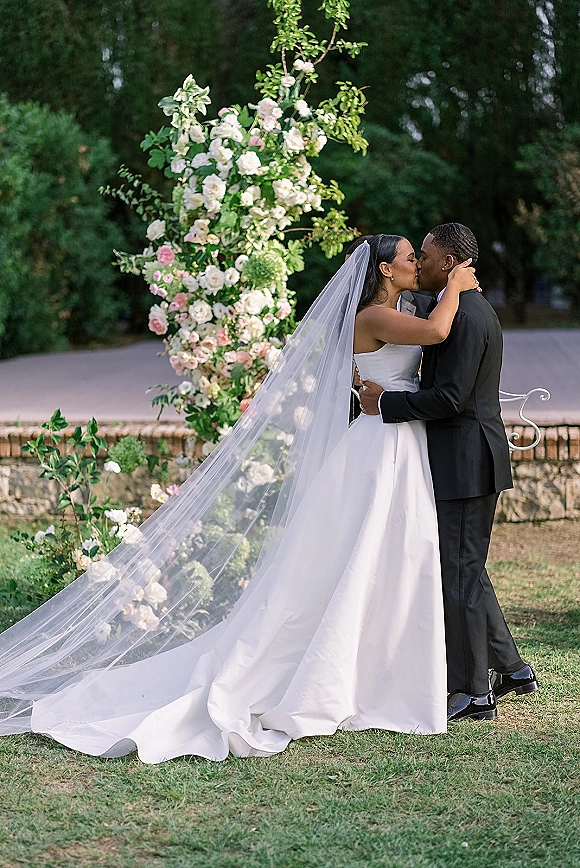 Wedding kiss portrait of bride and groom kiss under a rose and greenery floral arch, her long veil flowing in a garden setting