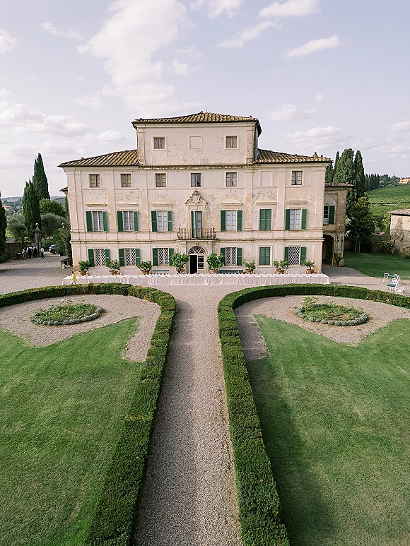 Wedding venue exterior with a long banquet table in white linens and chairs in a gravel courtyard before a historic villa with green shutters