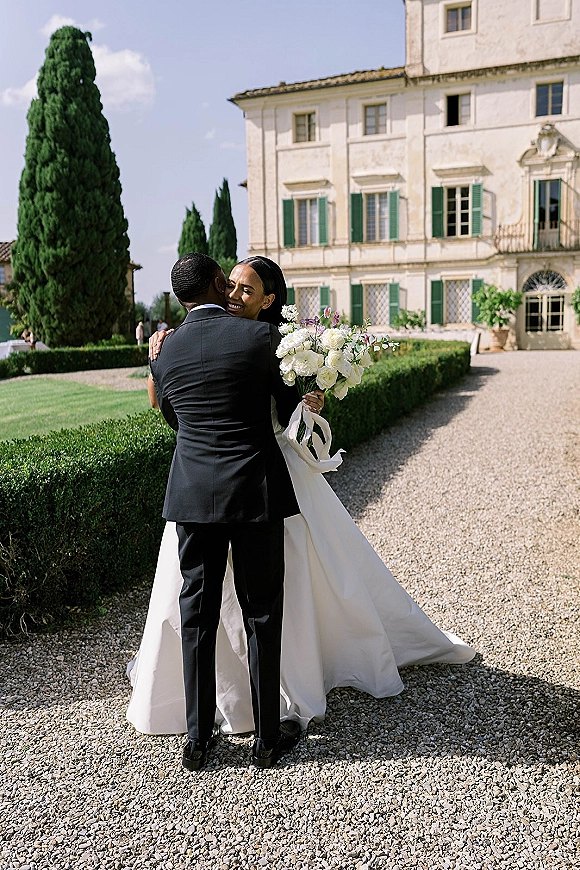 Couple portrait of bride and groom hug, bride holding a white bouquet, by a villa with green shutters, gravel drive, and cypress trees