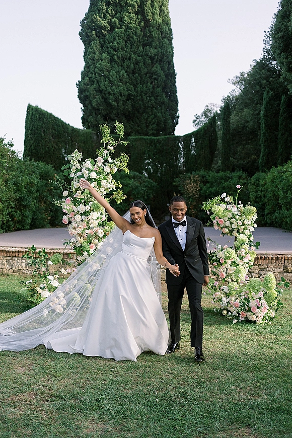 Couple portrait of bride and groom holding hands under a pastel floral arch, her long veil trailing, on a garden lawn with tall trees