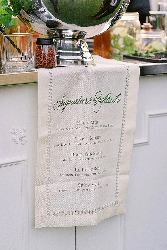 Signature cocktail menu on printed linen with calligraphy beside a silver beverage dispenser, rocks glass, and bar tools on a white bar counter