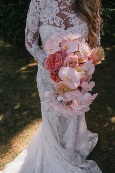 Bridal bouquet, cascading bridal bouquet of pink orchids, roses, and blush dahlias held against a long-sleeve lace dress in sunlit garden greenery