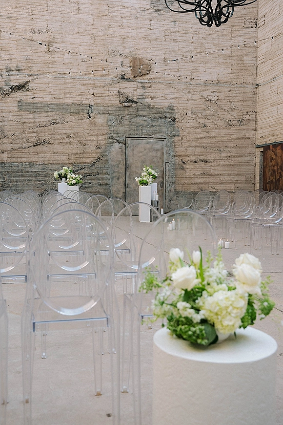 Ceremony setup for an indoor wedding ceremony with clear acrylic chairs, white floral plinths, and pillar candles against an industrial brick wall