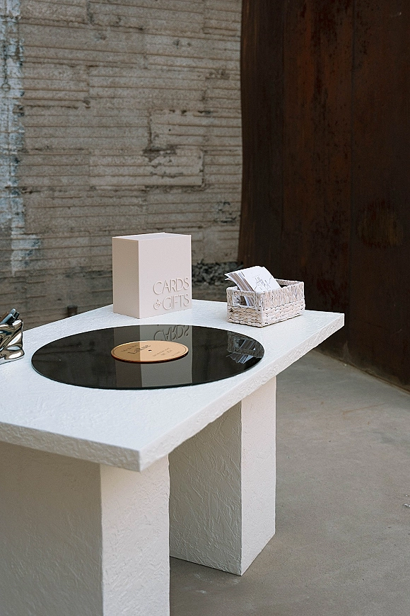 Wedding card table with cards and gifts box, wicker basket, and vinyl record turntable on a white pedestal against a concrete wall