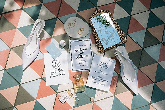Wedding invitation suite wedding flat lay with RSVP and details cards, white slingback heels, gold rings on colorful geometric tile in sunlight
