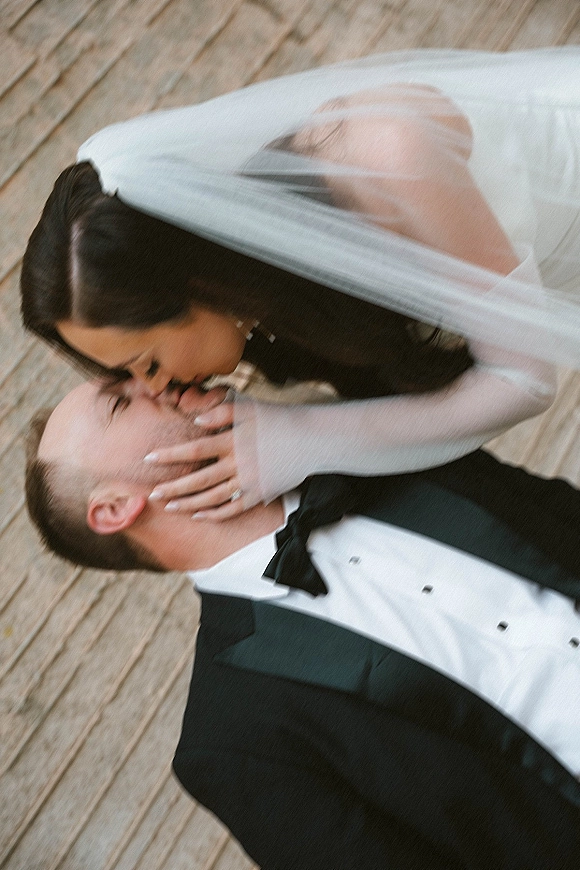 Wedding kiss portrait of bride kissing groom, veil over couple as she holds his face, tuxedo and rings visible on a wooden deck