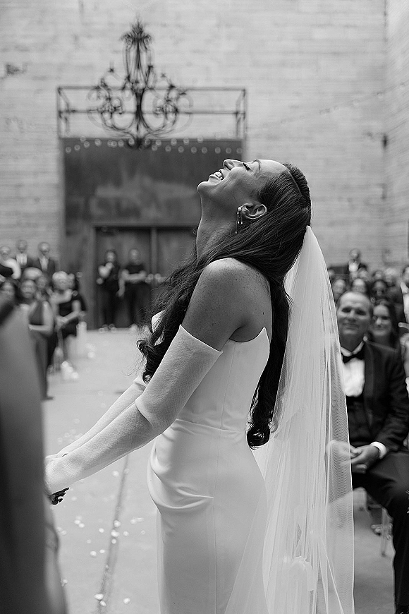 Bride portrait in a black and white wedding portrait, laughing in a veil with guests lining the aisle beneath a chandelier by brick wall