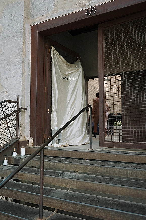 Wedding entrance decor with fabric draping and a custom monogram backdrop, candlelit on concrete steps at an industrial brick doorway