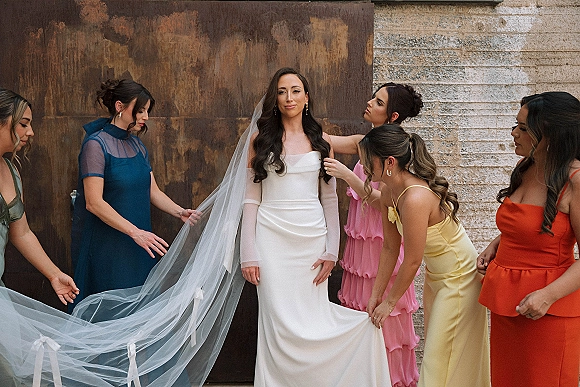 Bridal party portrait of bride with bridesmaids adjusting her cathedral veil, colorful dresses against a rust metal and concrete wall backdrop