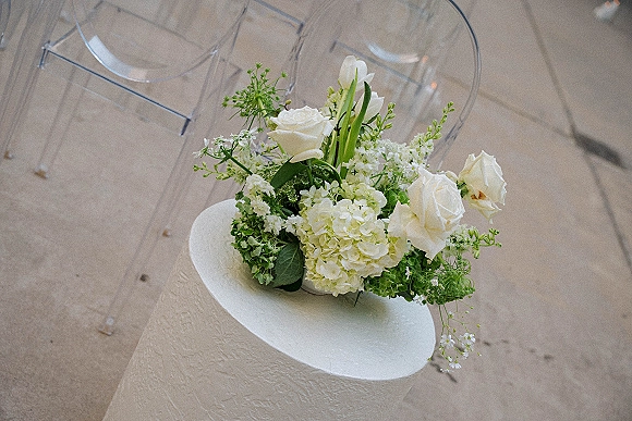Wedding cake with white wedding cake buttercream, adorned with roses and hydrangeas, on a pedestal beside clear acrylic chairs on wood floor