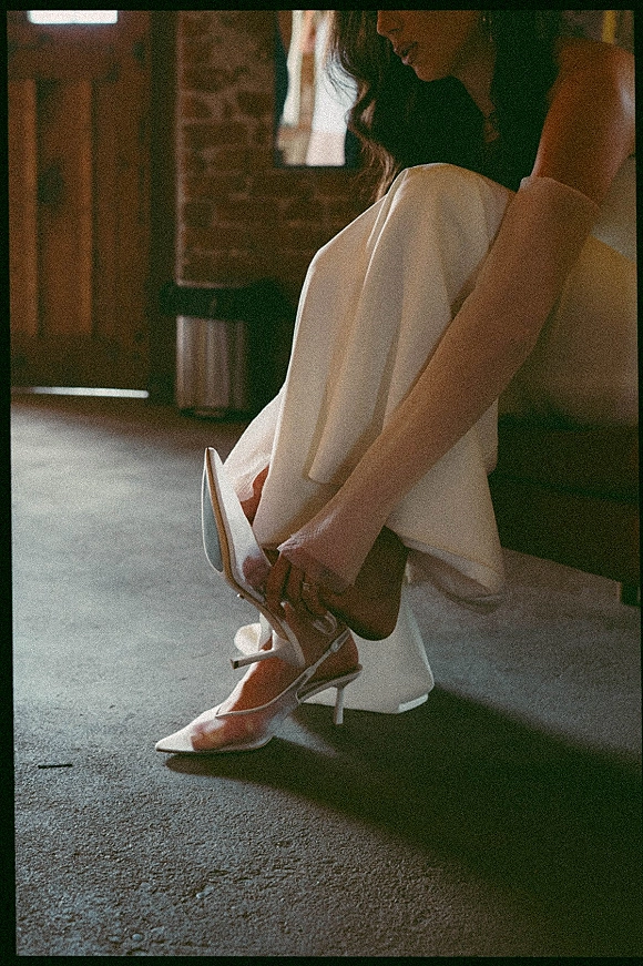 Bridal shoes in white ankle strap heels beside a wedding dress hem, engagement ring accent, set on an indoor floor by brick wall and door