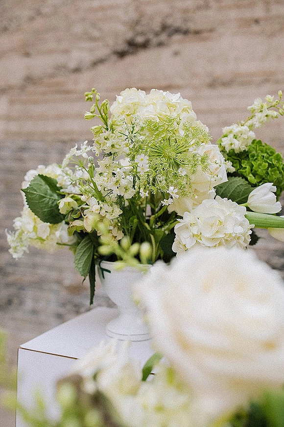 Wedding floral arrangement of white hydrangeas, roses and tulips with green foliage in a white pedestal vase against a stone wall outdoors