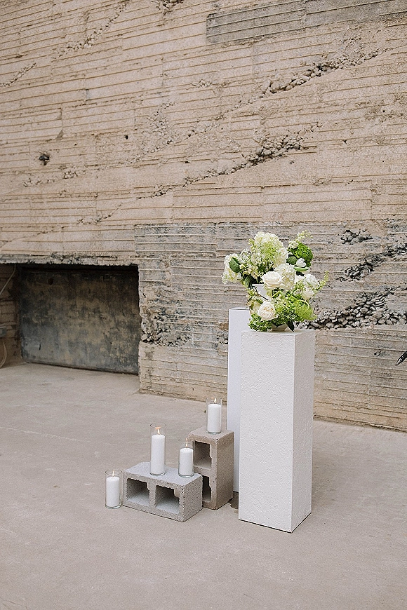 Ceremony altar decor with a modern wedding altar of white flowers and greenery on plinths, pillar candles and cinder blocks against concrete wall