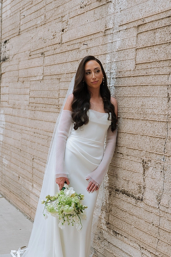 Bridal portrait of a bride in an off the shoulder wedding dress with veil, holding a bouquet beside a textured brick wall sidewalk