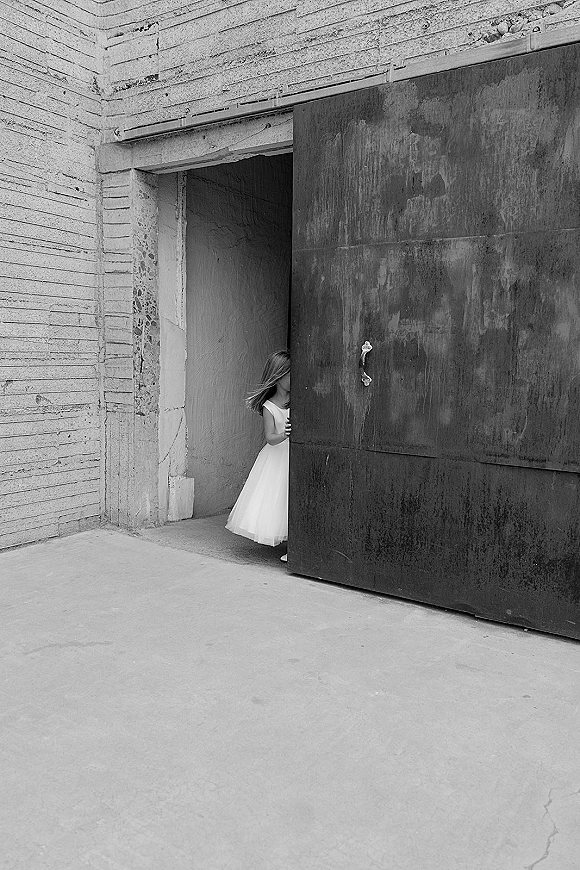 Bridal portrait of a bride peeking behind a metal door in a short wedding dress, framed by an industrial concrete doorway in black and white