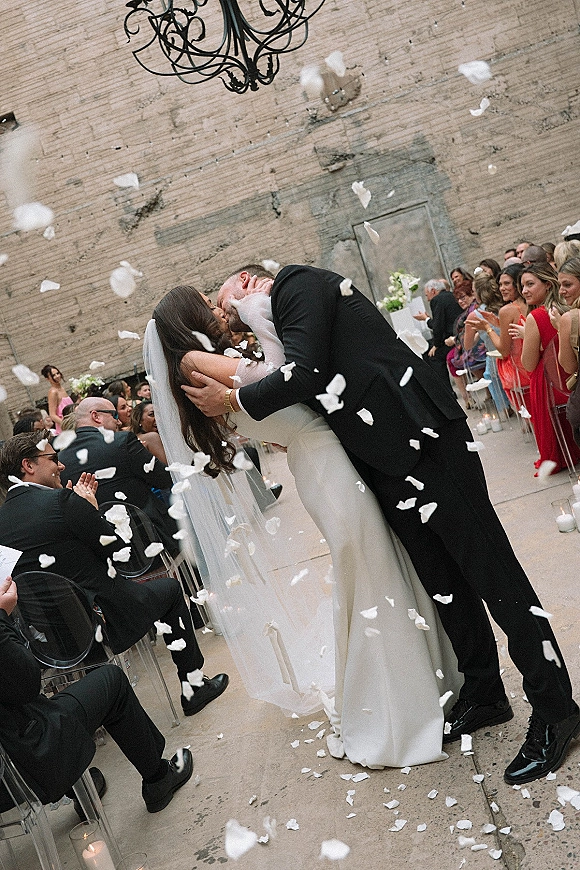 Wedding kiss as rose petals fall while bride in strapless dress and veil kisses groom in tux on candlelit aisle by concrete wall