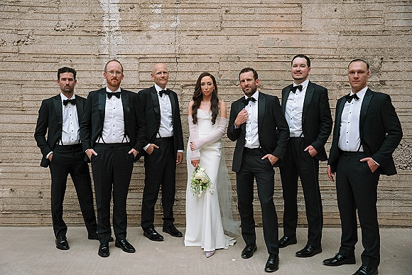 Wedding party portrait of a bride with groomsmen in black tuxedos and suspenders, holding a white bouquet against a textured concrete wall