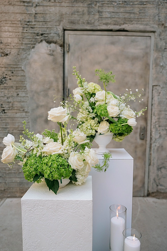 Wedding floral arrangement of white and green wedding flowers with roses, hydrangea and tulips on white plinths, candles by a concrete wall