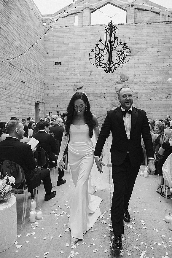 Wedding recessional as newlyweds walk down the aisle holding hands, candle-lined runner with rose petals under string lights in a brick-walled courtyard