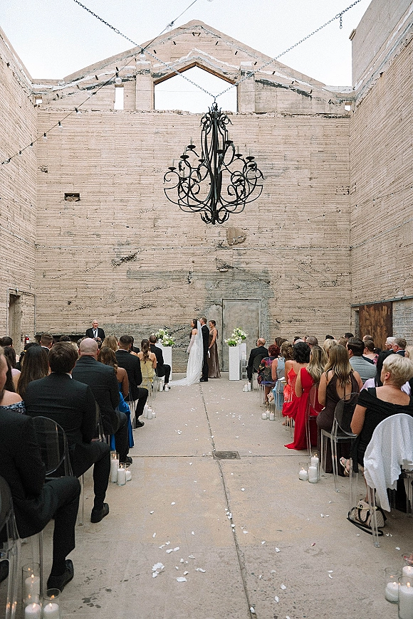 Wedding ceremony with bride and groom exchanging vows under chandelier and string lights, candles and petals lining a concrete aisle in an industrial venue