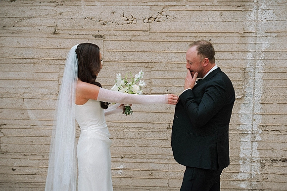 Wedding first look as bride taps groom’s shoulder, holding a white bouquet; groom in tux covers his mouth by a concrete wall