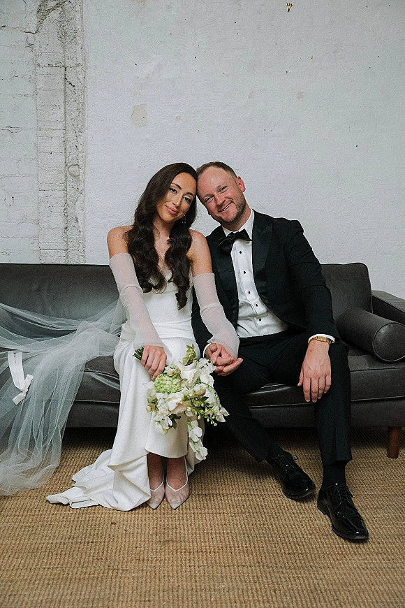 Couple portrait of bride and groom sitting on sofa, holding hands; long veil and bouquet against white plaster wall and exposed brick