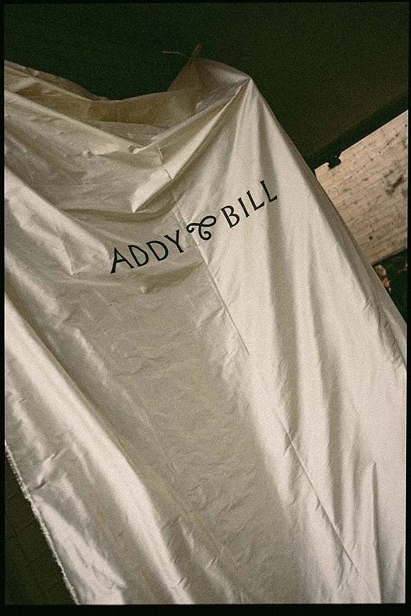 Wedding dress garment bag hanging with printed names lettering against a dark wall and door frame, ready for bridal gown transport