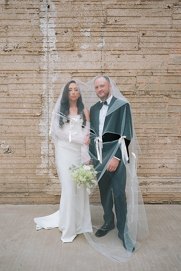Couple portrait of bride and groom portrait with a bridal veil draped over them, holding a white and green bouquet against a concrete wall