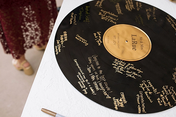 Wedding guest book vinyl record with gold lettering and handwritten messages on a white tablecloth, guests’ shoes visible nearby