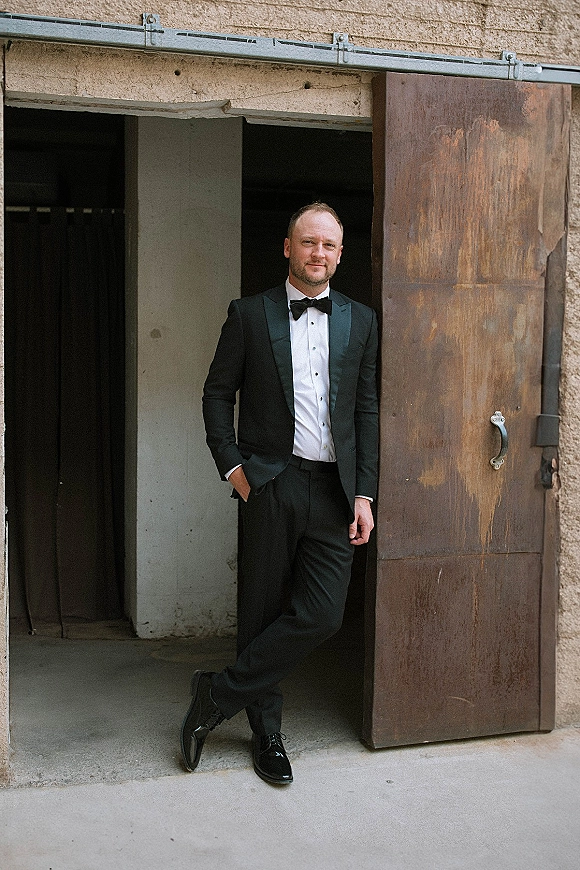 Groom portrait in a black tuxedo groom look with bow tie and hands in pockets, posed by an industrial doorway with metal door backdrop