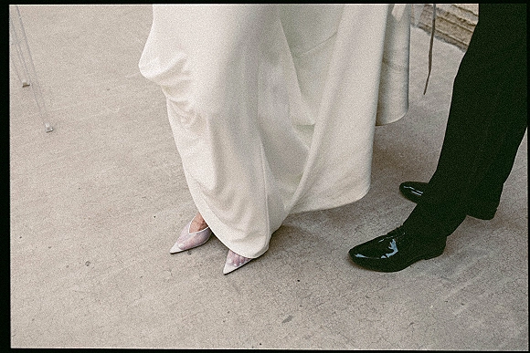 Wedding shoes beside groom black dress shoes on a concrete floor, featuring pointed toe heels under white wide leg pants