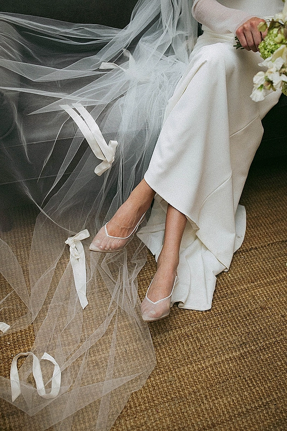 Bridal veil cathedral length veil draped on a carpeted floor with ribbon ties beside sheer wedding shoes, bouquet, and sofa