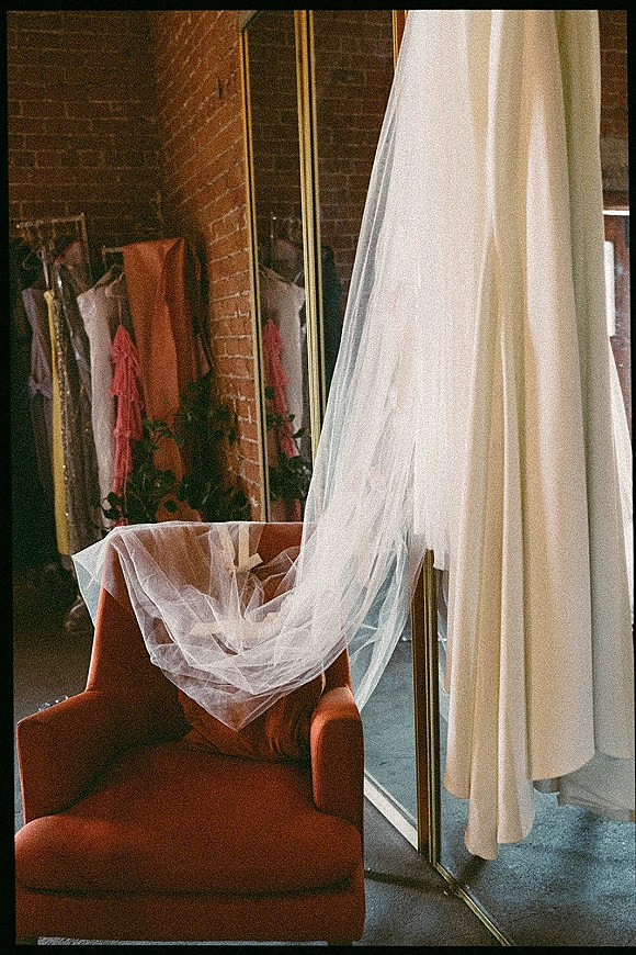 Wedding dress hanging on a garment rack with a long veil draped over a blush armchair, lit by window light against a brick wall