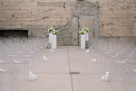 Ceremony aisle decor with clear acrylic ceremony chairs, a white runner lined with pillar candles and floral plinths against a concrete wall