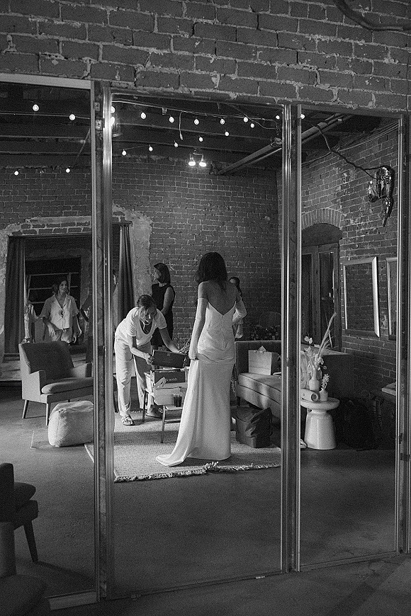 Getting ready moment as a bride steps into an off-the-shoulder wedding dress in a brick-walled lounge with string lights and dried florals