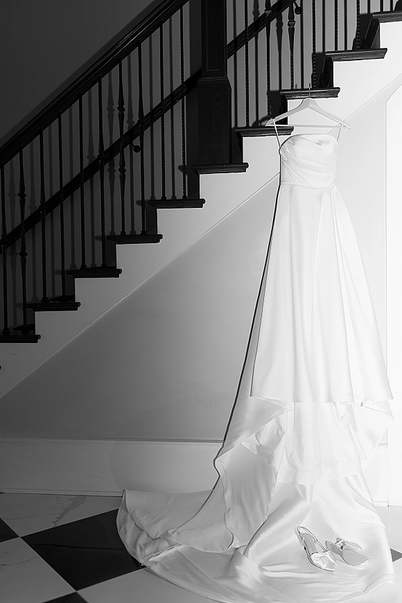 Wedding dress hanging on a staircase, strapless bridal gown with long train draped over checkered floor, bridal shoes nearby