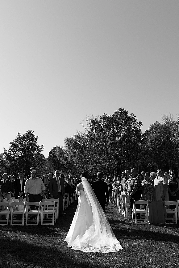 Wedding processional as bride walking down aisle in strapless wedding dress and long veil, guests standing beside white chairs on lawn