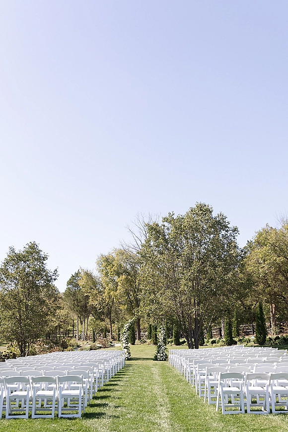 Ceremony setup for an outdoor wedding ceremony with white folding chairs lining an aisle to a round floral arch on a grassy lawn under blue sky