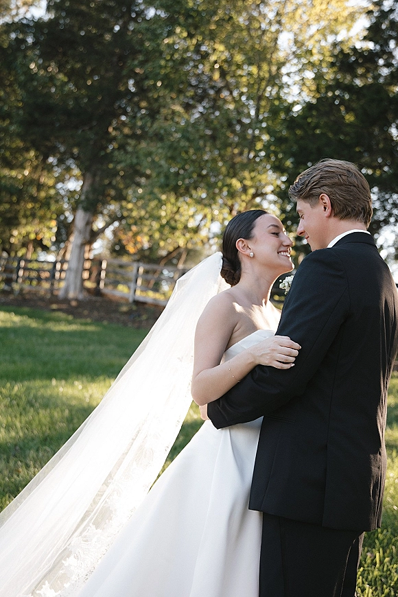 Wedding couple portrait of bride and groom embrace forehead to forehead, her long veil catching sunlight on a green lawn by trees and fence