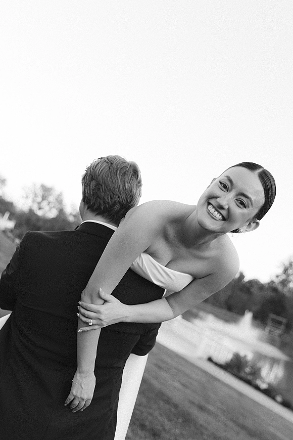 Couple portrait in a black and white wedding portrait, groom carrying bride piggyback as she laughs by a lakeside lawn with trees and sky