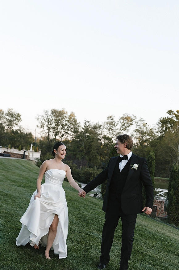 Couple portrait of bride and groom holding hands, walking on a grassy lawn near water and brick wall; bride in strapless gown, groom in tuxedo