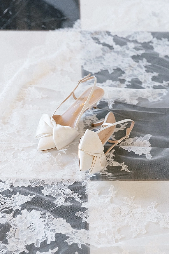 Bridal shoes with white bow wedding heels and ankle straps resting on a lace veil atop a soft fabric surface, pointed toes forward