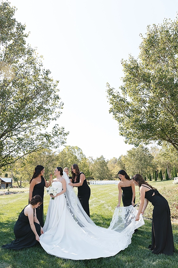Bride with bridesmaids helping bride adjust her strapless wedding dress and lace veil, holding a white rose bouquet on a lawn near ceremony chairs