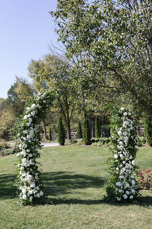 Wedding ceremony arch with white flowers and greenery on a grassy lawn, set before trees and a wooden fence under blue sky